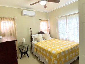 A bedroom with a bed, a ceiling fan, an air conditioner, a small table with a lamp, and curtains on the windows. - Cedarville, tranquil 2 bedroom house gated community Montegobay Jamaica  (Montego Bay)