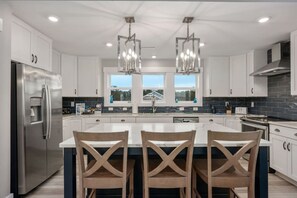 A modern kitchen with stainless steel appliances, a central island with bar stools, and pendant lighting. - Baydream Believer | Soundfront, Pool & Hot Tub (Corolla)