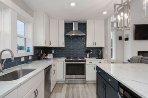 A modern kitchen with white cabinetry, a stainless steel range hood, and a black backsplash. - Baydream Believer | Soundfront, Pool & Hot Tub (Corolla)