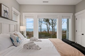 A bedroom with a large bed, a view of the beach, and a door leading outside. - Baydream Believer | Soundfront, Dock, Heated Pool & Hot Tub (Corolla)