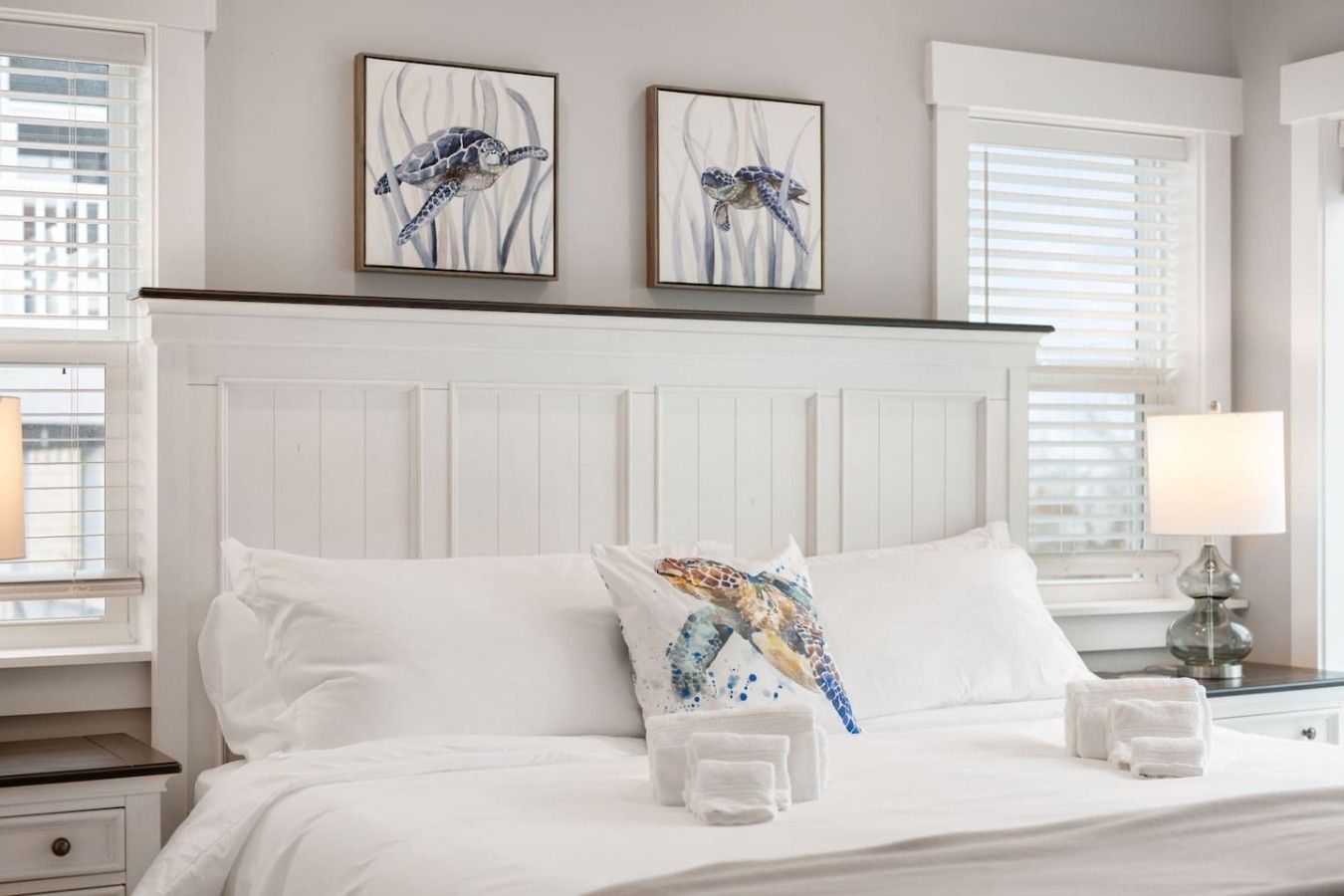 A hotel room with a large bed, white bedding, two wall-mounted paintings of sea turtles, a bedside lamp, and a window with blinds.