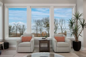 A living room with two armchairs, a small table, and a large window with a view of trees and a fence. - Baydream Believer | Soundfront, Pool & Hot Tub (Corolla)