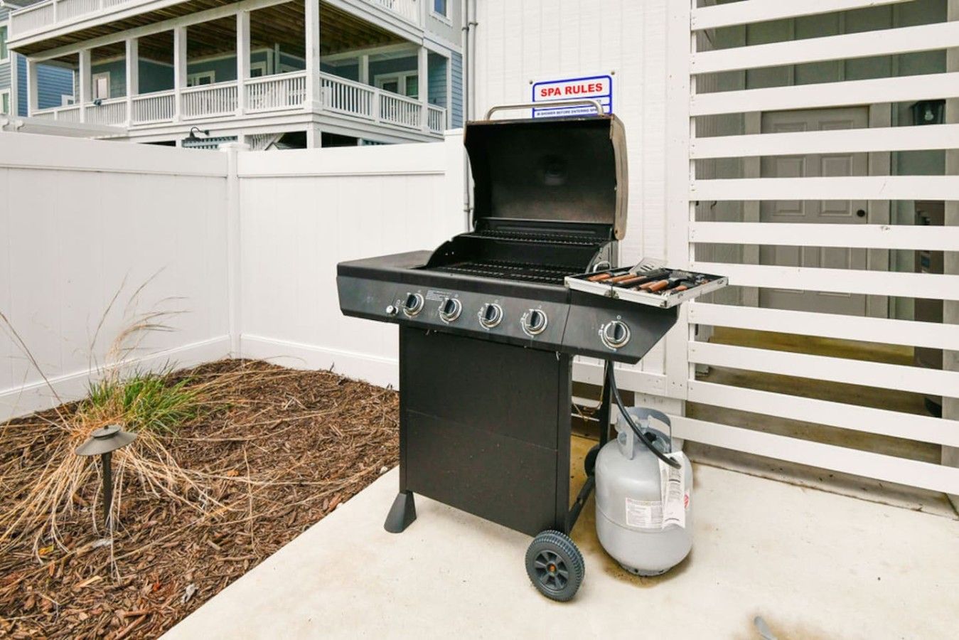 A portable gas grill with a sign indicating 'SPA RULES' on a white fence.