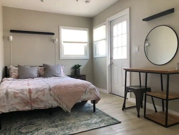 Cozy retreat with a comfortable bed, bedside table, and potted plant, plus a large mirror and window with blinds.