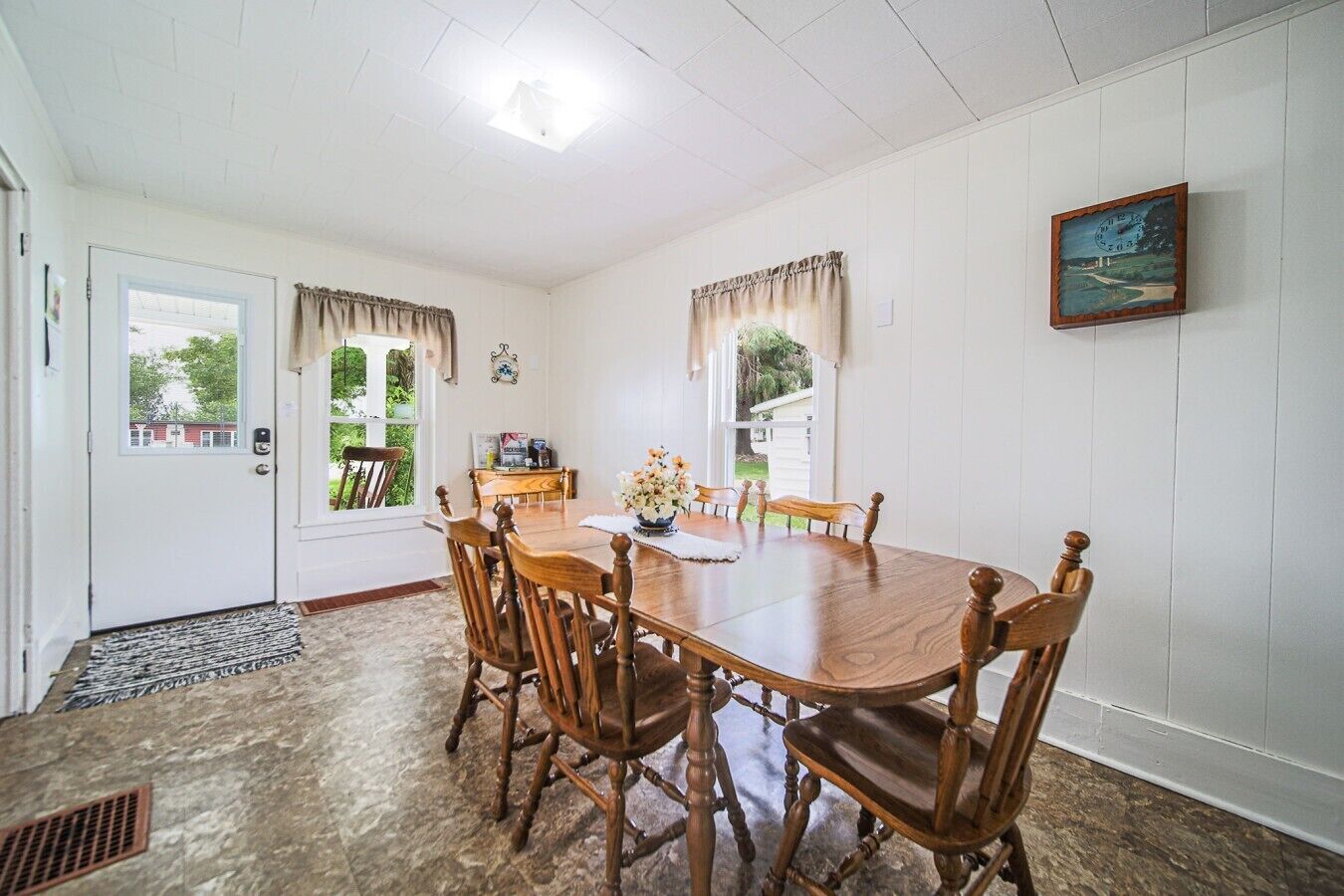 Une salle à manger avec une table et des chaises en bois, une horloge au mur et une fenêtre avec des rideaux.
