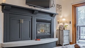 A living room with a dark grey built-in entertainment unit, a fireplace, a television mounted above, and a small cabinet with a lamp. - Basecamp Stays: Upscale Couple's Getaway, Hot Tubs (Driggs)