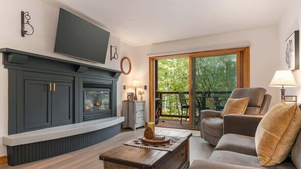 A living room with a fireplace, a sofa, a coffee table, and a large window with a view of trees. - Basecamp Stays: Upscale Couple's Getaway, Hot Tubs (Driggs)