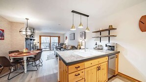 A modern kitchen with wooden cabinets, a central island, pendant lighting, and a dining area with a table and chairs. - Basecamp Stays: Upscale Couple's Getaway, Hot Tubs (Driggs)