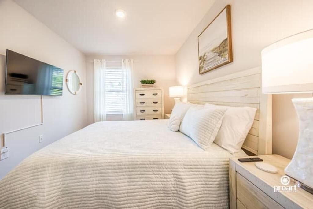 Un dormitorio ordenado con cama, mesita de noche, lámpara y televisor montado en la pared.