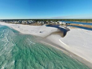 Vista aérea de uma praia com águas turquesa cristalinas, uma faixa de areia e uma fileira de casas ao fundo.