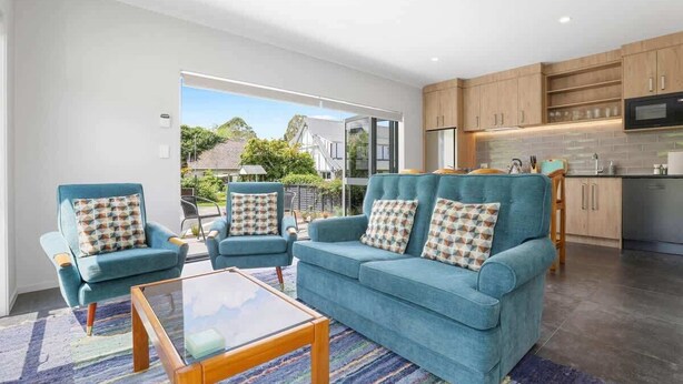 A modern living room with a blue sofa and armchairs, a glass coffee table, and a kitchen area with wooden cabinets and a microwave. - All Good - MTB Base, Bike Wash, Sleeps 4 (Rotorua)