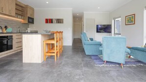 A modern kitchen with wooden cabinets, a dining area with a wooden table and chairs, a blue sofa, and a flat-screen TV in the living room. - All Good - MTB Base, Bike Wash, Sleeps 4 (Rotorua)