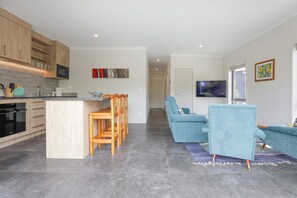 A modern kitchen with a central island, wooden chairs, a blue sofa, and a flat-screen TV. - All Good - MTB Base, Bike Wash, Sleeps 4 (Rotorua)
