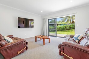 A living room with two leather sofas, a flat-screen TV mounted on the wall, a small wooden coffee table, and a sliding glass door leading to a garden.