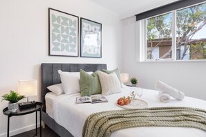A neatly made bed with pillows, a bedside table with a lamp, a framed picture on the wall, and a window with a view.