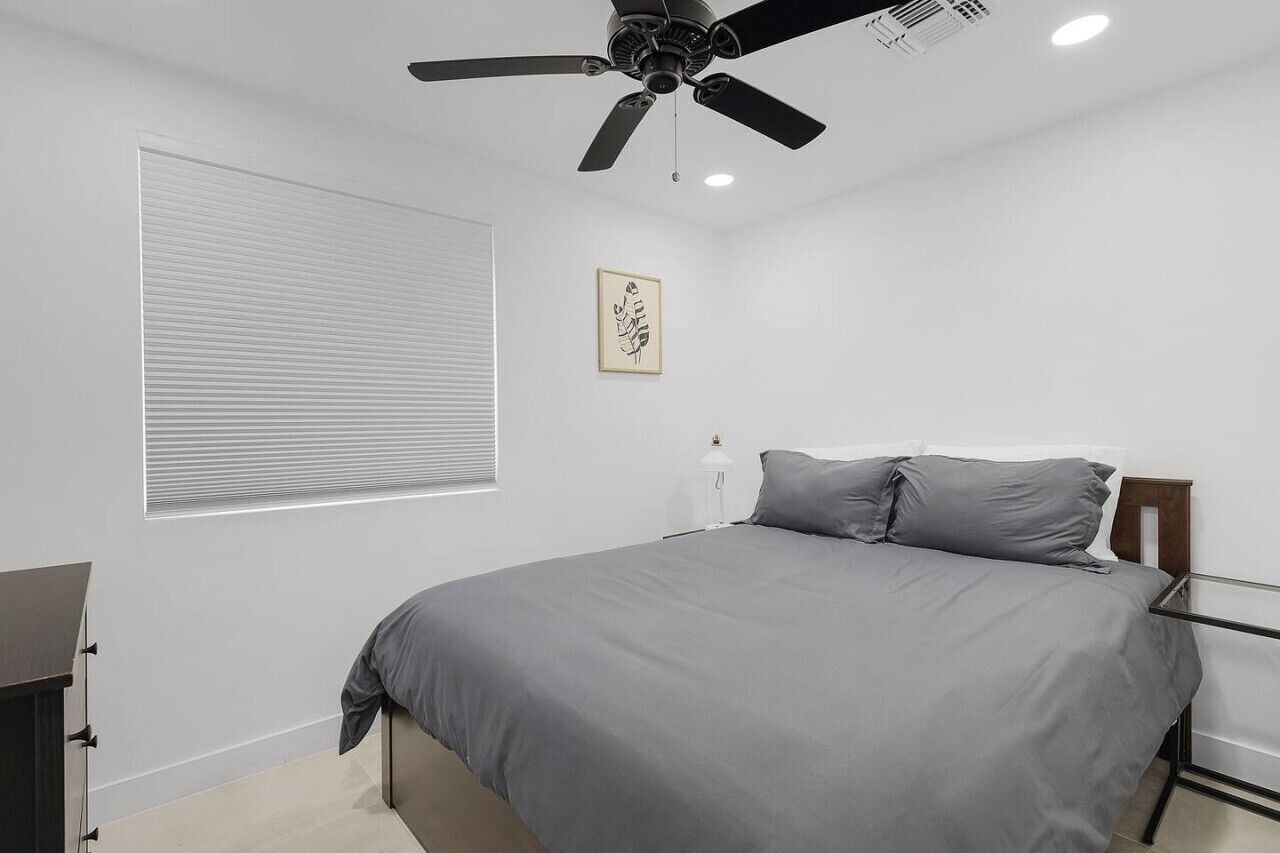 Un dormitorio con una cama, un ventilador de techo, una ventana con persianas y una obra de arte enmarcada en la pared.