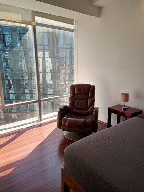 A bedroom with a large window, a brown recliner, a bedside table with a lamp, and a bed.