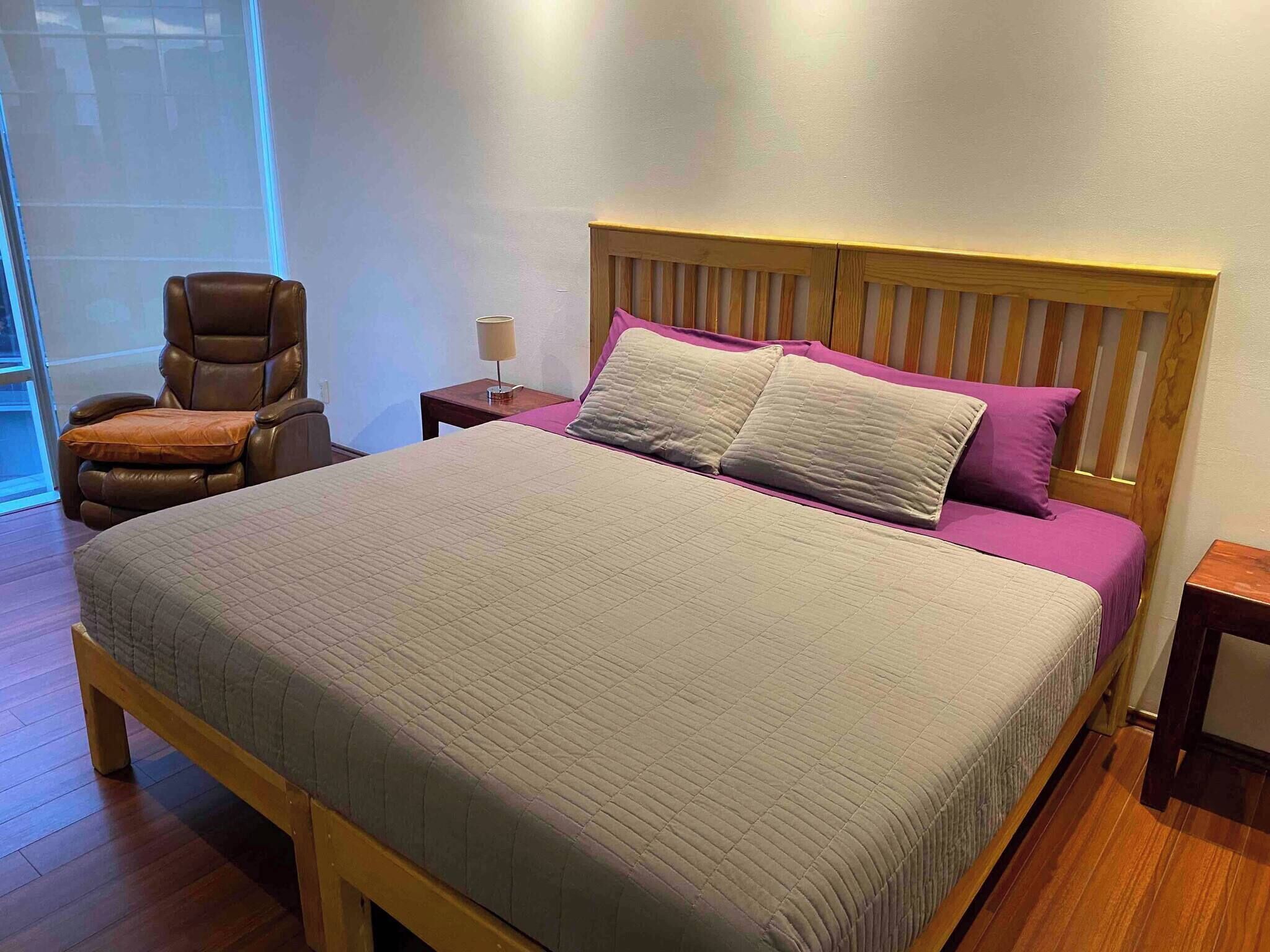 Un lit avec une tête de lit en bois, un fauteuil en cuir marron, une table de chevet avec une lampe et une fenêtre avec des stores.