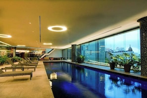 An indoor swimming pool with lounge chairs, a staircase, and a view of a cityscape through large windows.