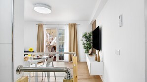 A modern hotel room with a wooden floor, a flat-screen TV mounted on the wall, a dining area with a table and chairs, and a large window with curtains. - Holiday Apartment 'Liomor Luxury' with Shared Pool, Wi-Fi and Air Conditioning (Caleta de Fuste)