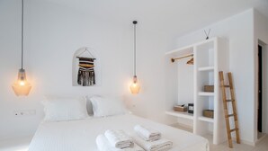 Una cama bien hecha con toallas enrolladas, un perchero y una estantería blanca con cestas.