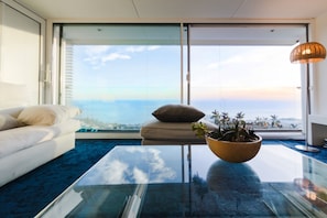 Een moderne woonkamer met een glazen salontafel, een witte bank, een beige kussen en uitzicht op zee.