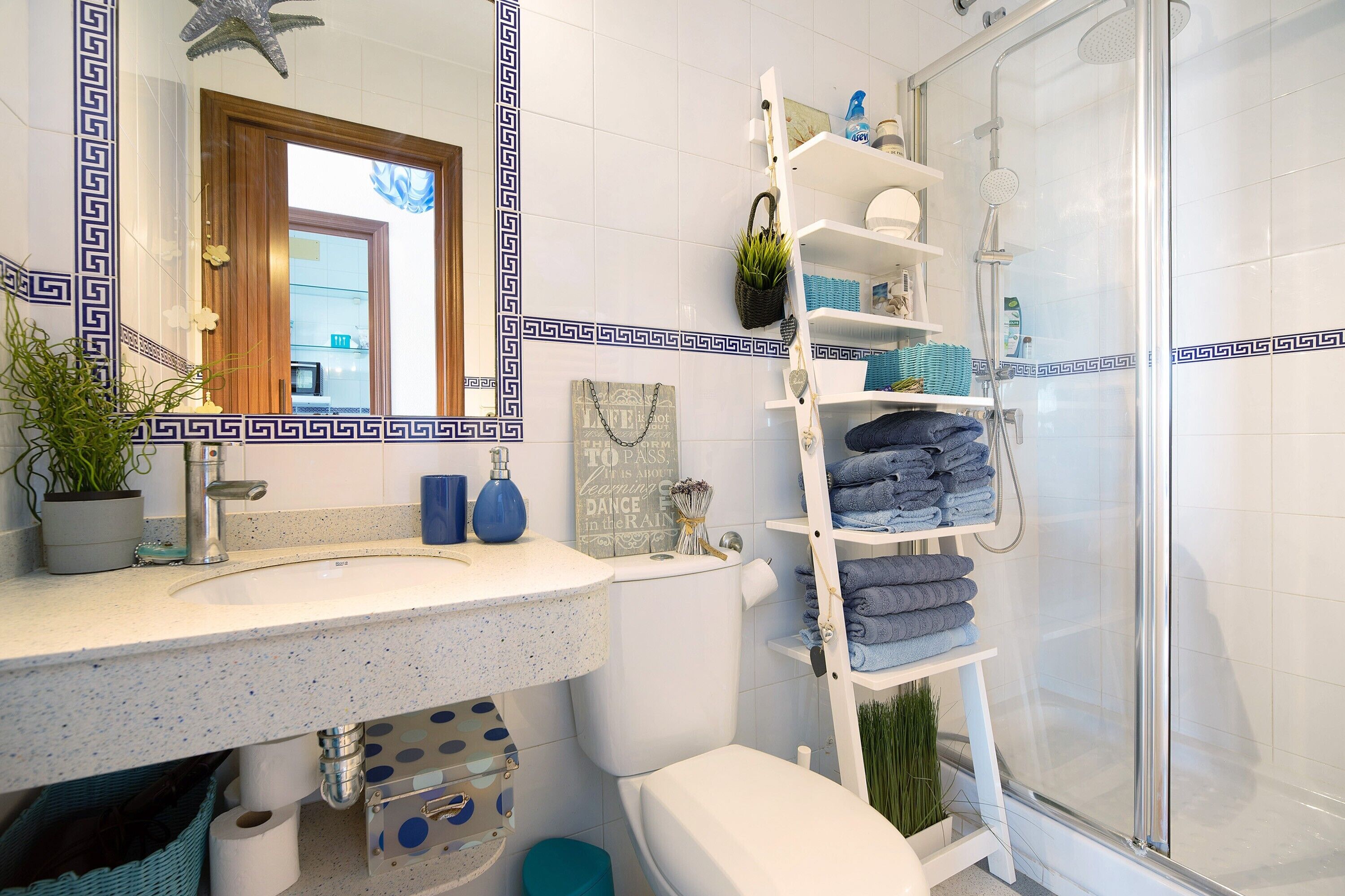 Un baño con lavamanos, inodoro y ducha blancos. Se aprecia un estante con toallas y una planta en maceta.