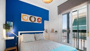 A bedroom with a blue wall, a bed with patterned bedding, a bedside table with a lamp, and artwork. - Apartment 'La Barca' with Shared Terrace, Wi-Fi, and Air Conditioning (Chipiona)