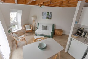Una habitación compacta con cama, una mesa de comedor con un tazón, sillas de mimbre, una kitchenette pequeña y un ventanal.