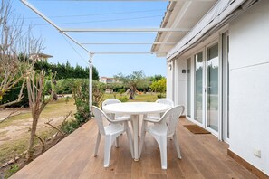 En patio med et hvidt bord og stole, et pergola og udsigt over en have samt tilstødende bygninger.