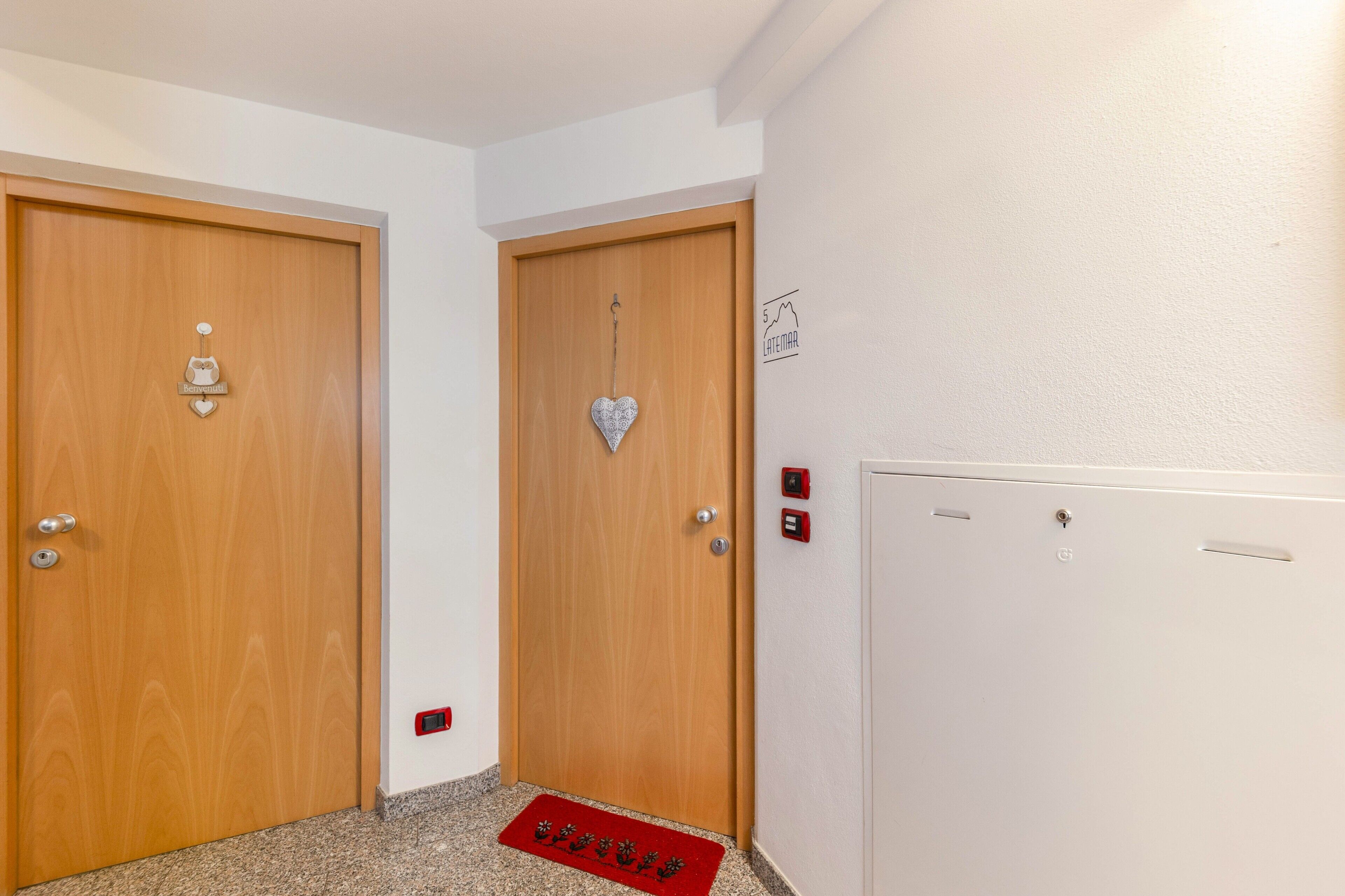 Un couloir avec deux portes en bois, un paillasson rouge et un mur blanc sur lequel est affiché un panneau.