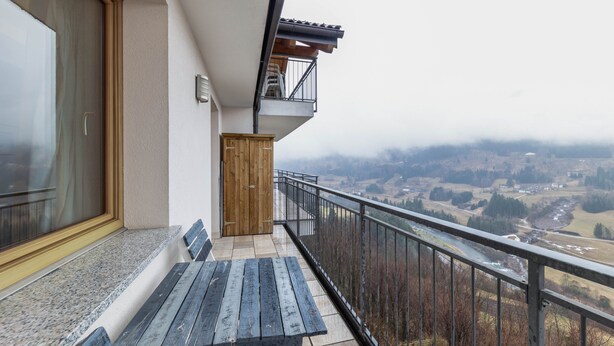 Een balkon met een houten bankje, uitzicht op een vallei en een metalen hekwerk.