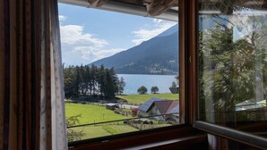 A view through a window with a wooden ceiling, a mountain, a lake, and a green landscape. - Holiday Apartment 'Appartements Egart 2' with Lake View, Balcony and Wi-Fi (Reschen)