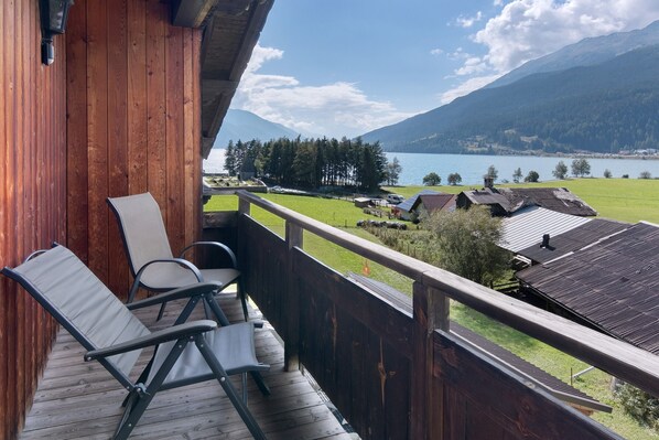 A wooden deck with chairs, a view of a lake, mountains, and a village.