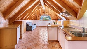 Een zolderkamer met hoge plafonds, een keuken, een eethoek en een raam met uitzicht op groen.