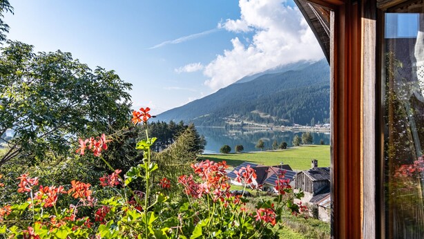 Uitzicht vanuit een raam op een meer, bergen en een landelijk gebied met traditionele gebouwen.