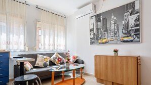 Uma sala de estar moderna com uma mesa de centro de vidro, um sofá com almofadas estampadas, um armário de madeira e uma pintura com vista para a cidade na parede.