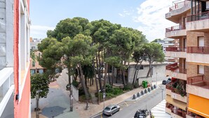 Um edifício de vários andares com varandas, uma rua com carros e árvores ao fundo.