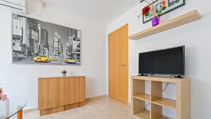 Quarto com armário de madeira, mesa de vidro, televisão sobre suporte de madeira, porta e pintura de paisagem urbana.