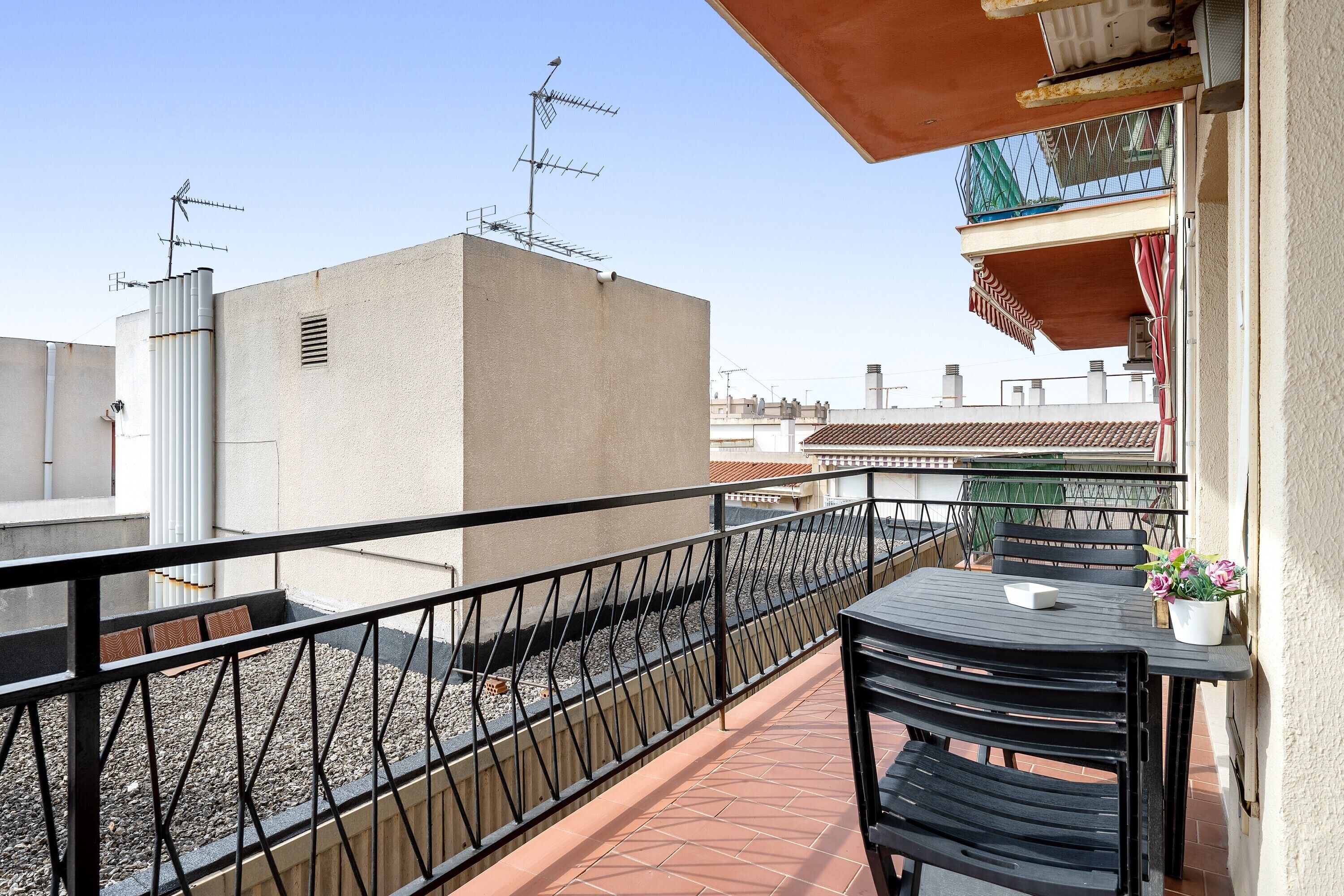 Un balcón con mesa y sillas, una barandilla y un edificio con antenas al fondo.