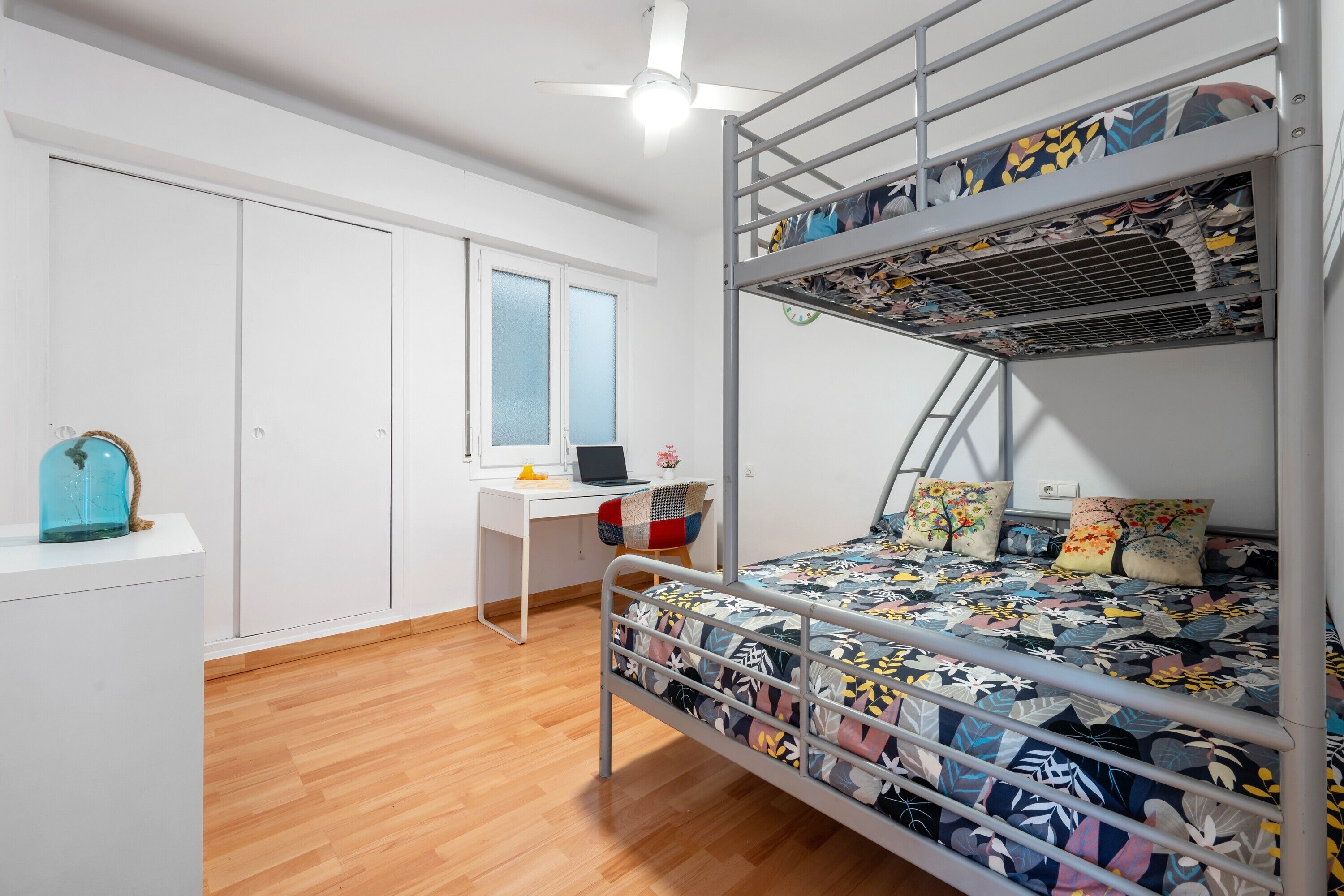 A bunk bed with a patterned bedspread, a desk with a laptop, a chair, and a window with blinds.