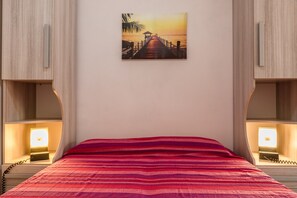 A hotel room with a bed, a painting of a sunset over a pier, and bedside lamps.