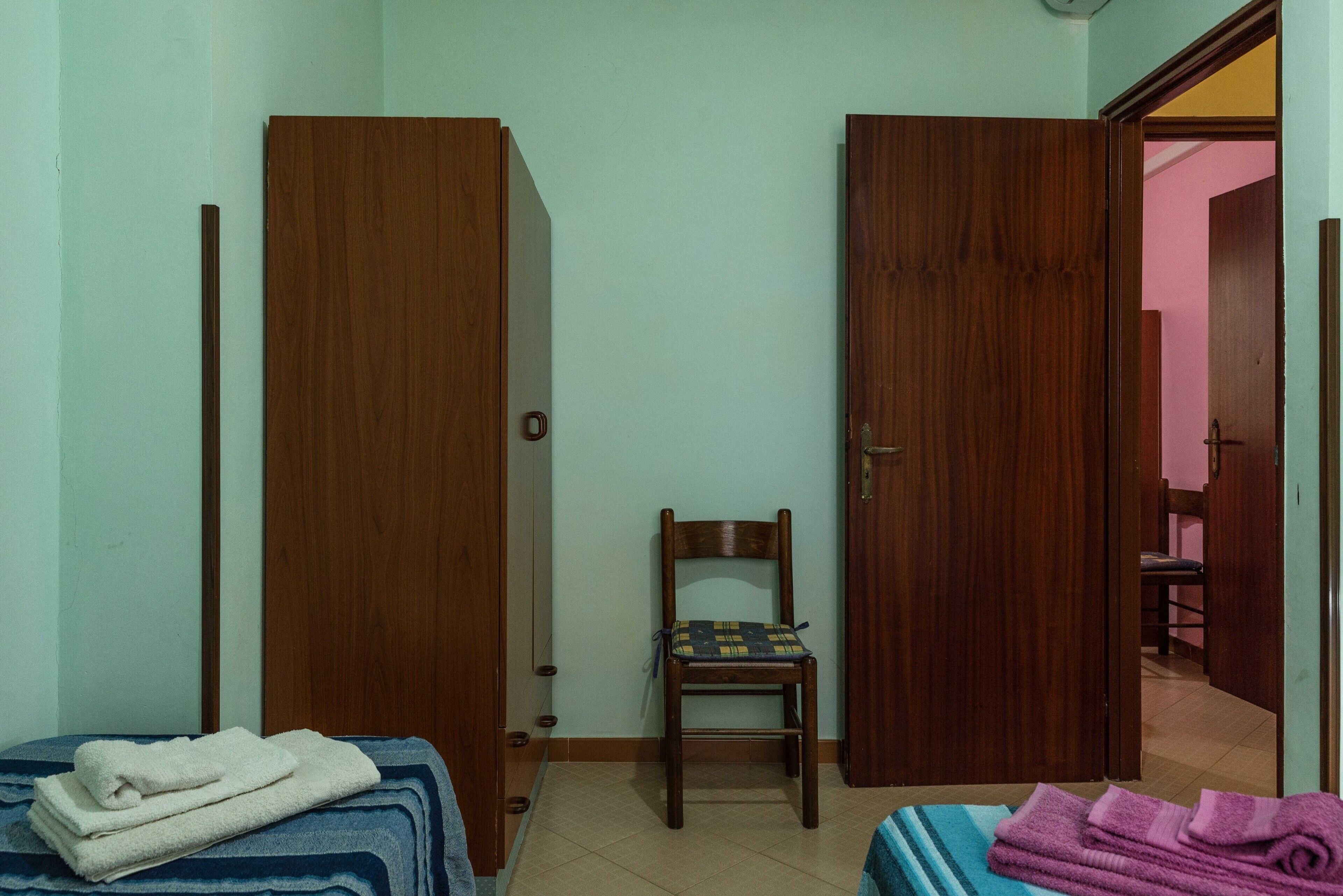 A room with two wooden doors, a chair, a bed with folded towels, and a visible bathroom in the background.