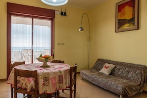 A dining area with a table set for two, a sofa, a window with a view, and a painting on the wall.
