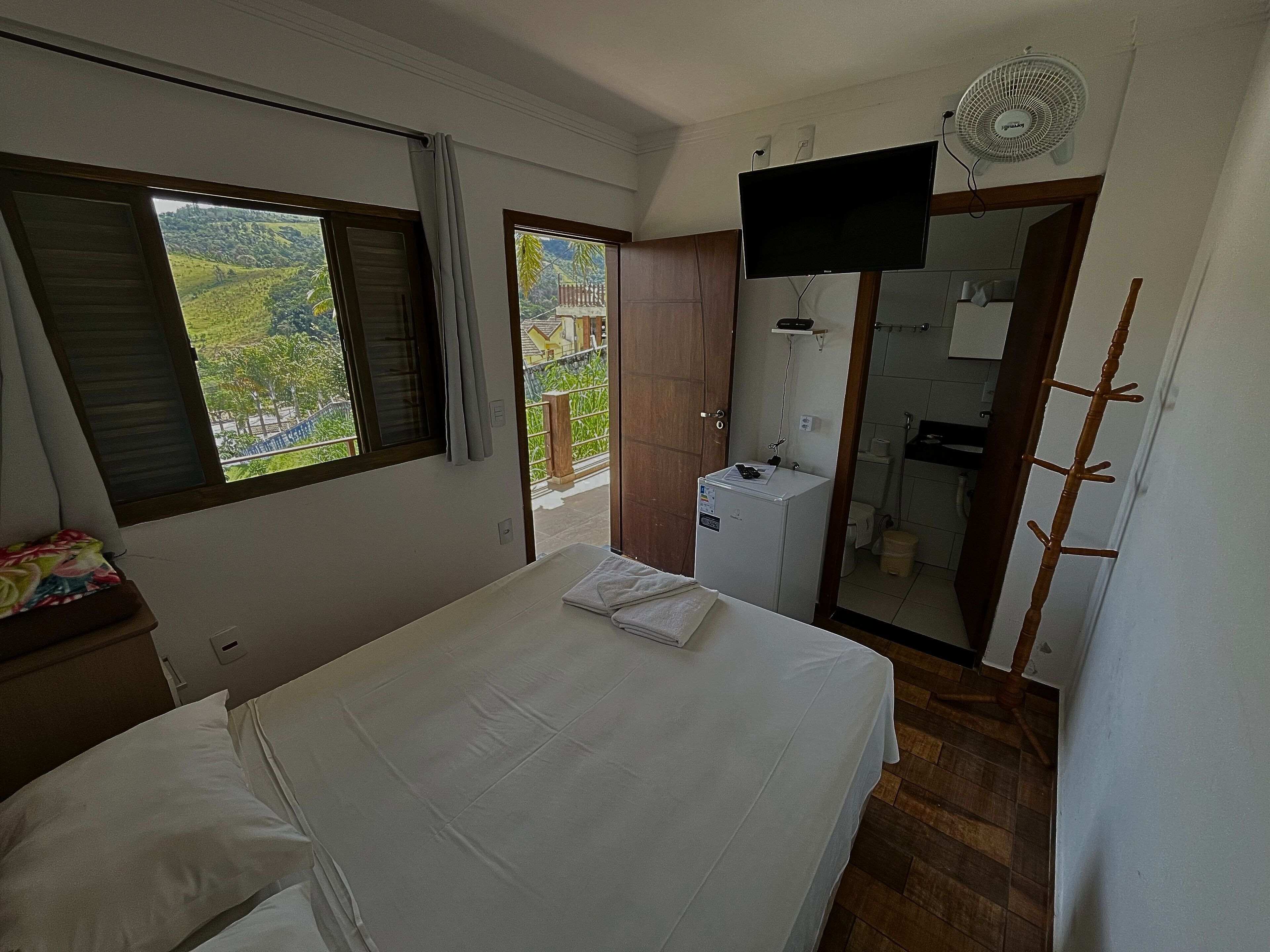 A bedroom with a bed, a small desk, a TV, a fan, and a bathroom visible through an open door.