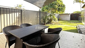 Un patio al aire libre con una mesa y sillas, una zona con sombra y vistas a un jardín trasero con valla y follaje.