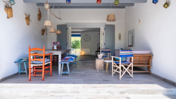 Un espace de vie à aire ouverte comprenant une table et des chaises, un banc en bois et une vue dégagée sur l’extérieur grâce à une porte ouverte.