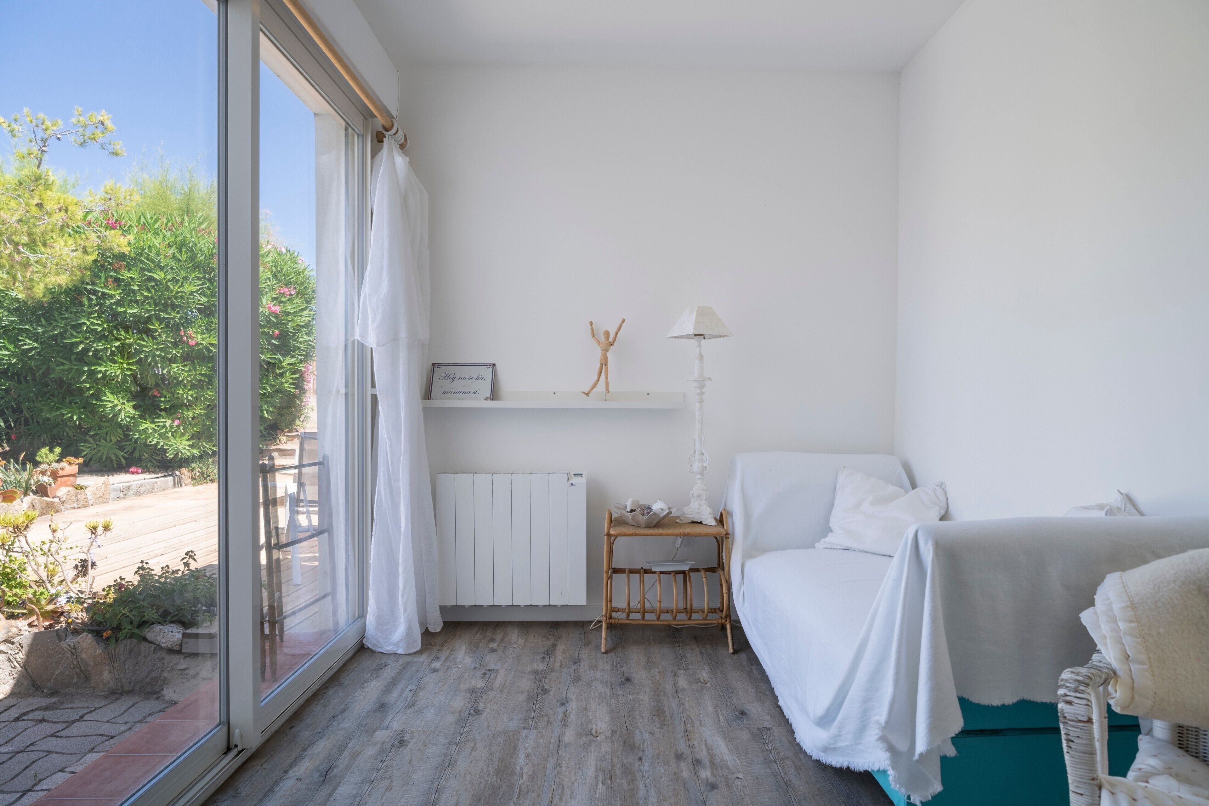 A bedroom with a large window, a bed, a bedside table with a lamp, and a small shelf with decorative items.