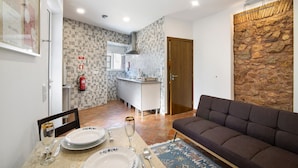Ein modernes Wohnzimmer mit einer Couch, einem Esstisch gedeckt mit Tellern, Gläsern und Besteck, einer Küche im Hintergrund und einer Wand mit Steinverkleidung.