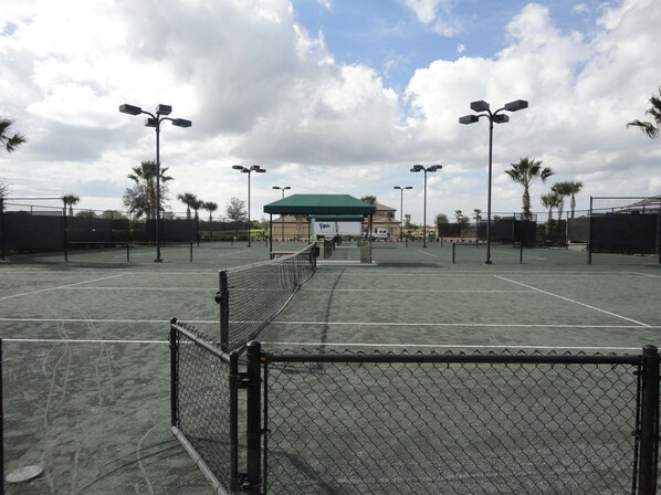 A tennis court with a chain-link fence, street lamps, and a green-roofed building in the background.